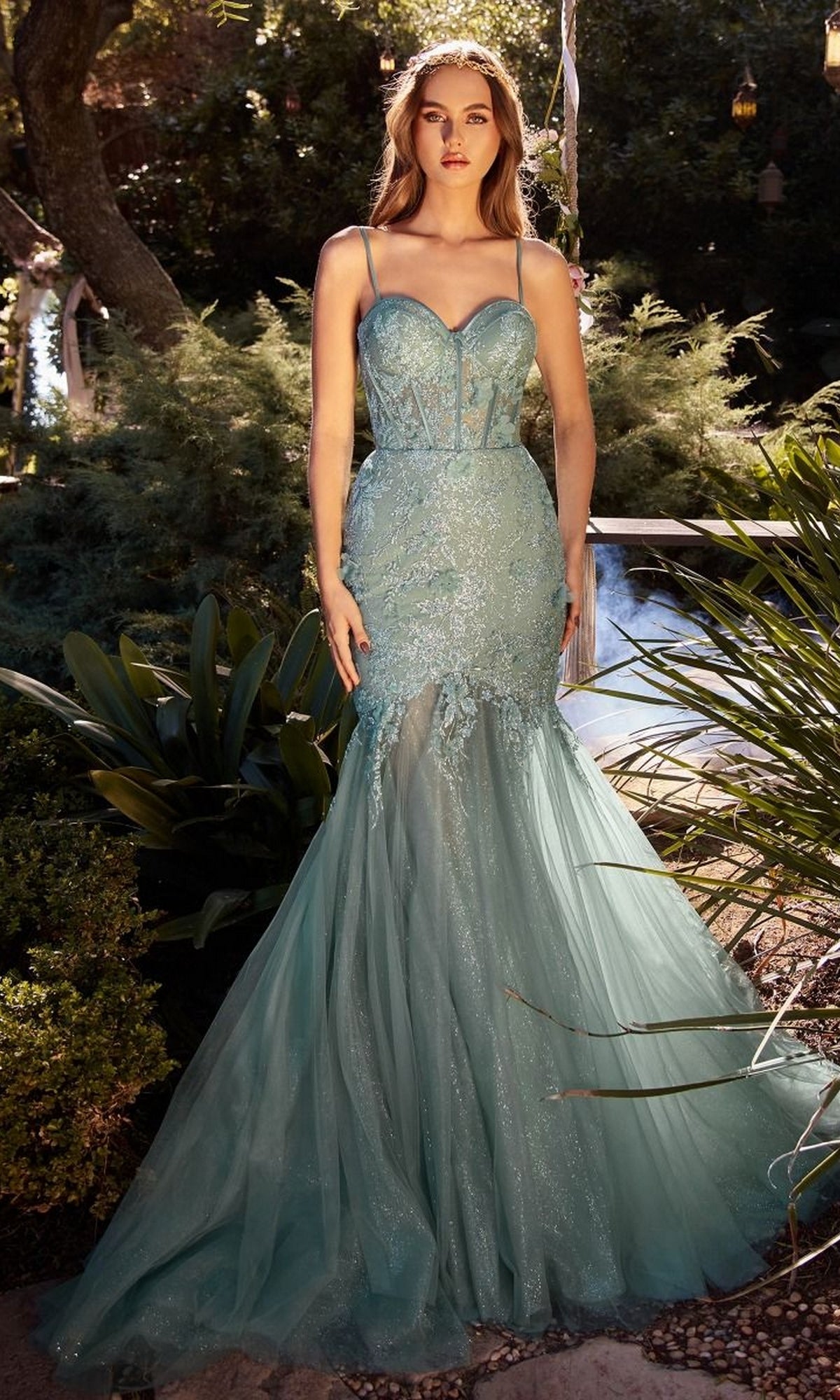 A woman is outdoors in the Andrea and Leo A1232 long lace mermaid prom dress, featuring a fitted light blue design with thin straps, intricate lace details, and a flowing tulle skirt.